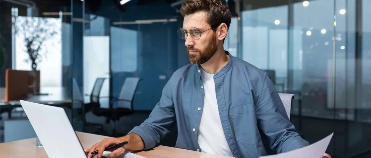 A man financier, auditor sits in the office . He holds documents, checks accounts. types on a laptop A serious young man accountant, financier, analyst, auditor sits in the office at the table. He holds documents and a pen in his hands, checks accounts, finances, types on a laptop.