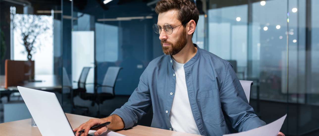 A serious young man accountant, financier, analyst, auditor sits in the office at the table. He holds documents and a pen in his hands, checks accounts, finances, types on a laptop.